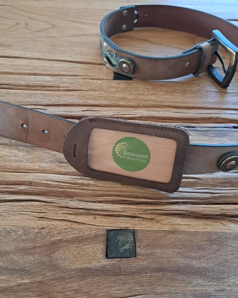 Leder-Anhänger mit Energiekarte BewusstPräsentSein, auf einen Ledergürtel gefädelt, auf einem Holztisch; dahinter ein Lederhalsband